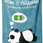 A blue-green Craftique throw blanket with "Mom is Charging" text, folded on a sofa, representing a cozy gift for moms.