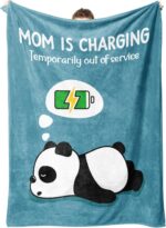 A blue-green Craftique throw blanket with "Mom is Charging" text, folded on a sofa, representing a cozy gift for moms.