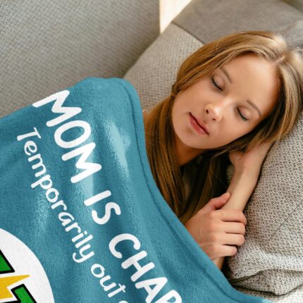 A blue-green Craftique throw blanket with "Mom is Charging" text, folded on a sofa, representing a cozy gift for moms.