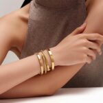 Two ISELBDR 18K gold plated stainless steel bangles, one with a clover design and one with a nail design, stacked together.