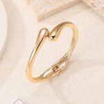 Four gold-plated bracelets, including a floral clover, tennis, chunky twist cuff, and classic bangle, stacked on a wrist.